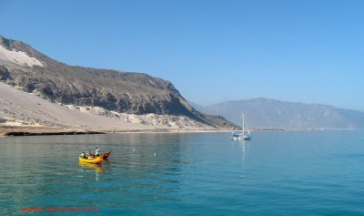 blue flat water with a little yellow fishing boat in the foreground and a yacht at anchor behind, with high stony mountains surrounding the bay