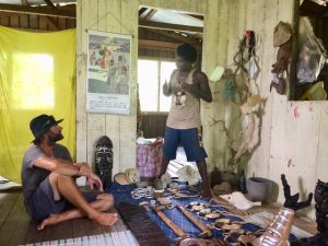 inside a wooden room with a local man standing surrounded by art and carvings and a crew sitting on the floor with a hat and shorts and t shirt listening